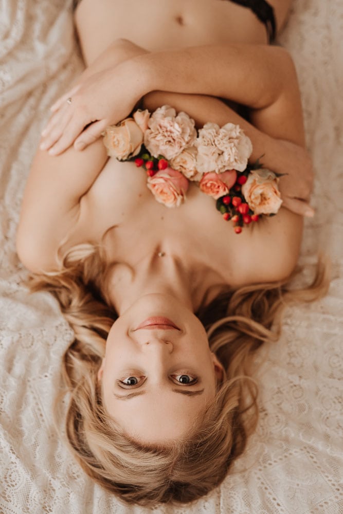Junge Frau liegt auf dem Bett, die Brüste mit Blumen verdeckt und schaut freundlich im die Kamera.